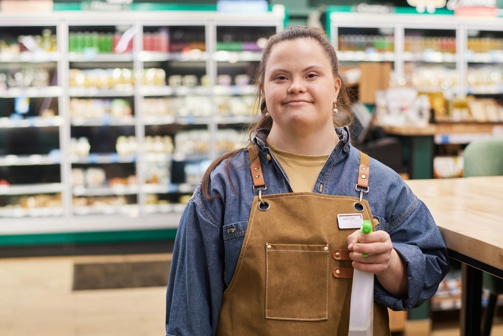 Waist up portrait of smiling young woman with Down syndrome working in supermarket and holding cleaning spray bottle copy space