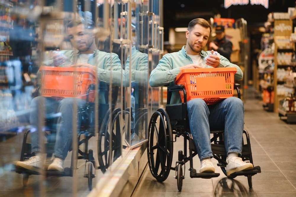 Man using wheelchair shopping groceries in supermarket, holding basket and choosing products from refrigerated shelves